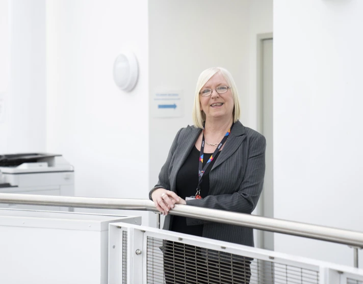 Staff member standing by a railing in a bright corridor at Glasgow Kelvin College, smiling at the camera Staff member standing by a railing in a bright corridor at Glasgow Kelvin College, smiling at the camera